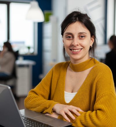 Close up of authentic professional employee looking at camera sitting at desk in front of laptop smiling, working financial startup company. Diverse team analyzes statistics data in modern office